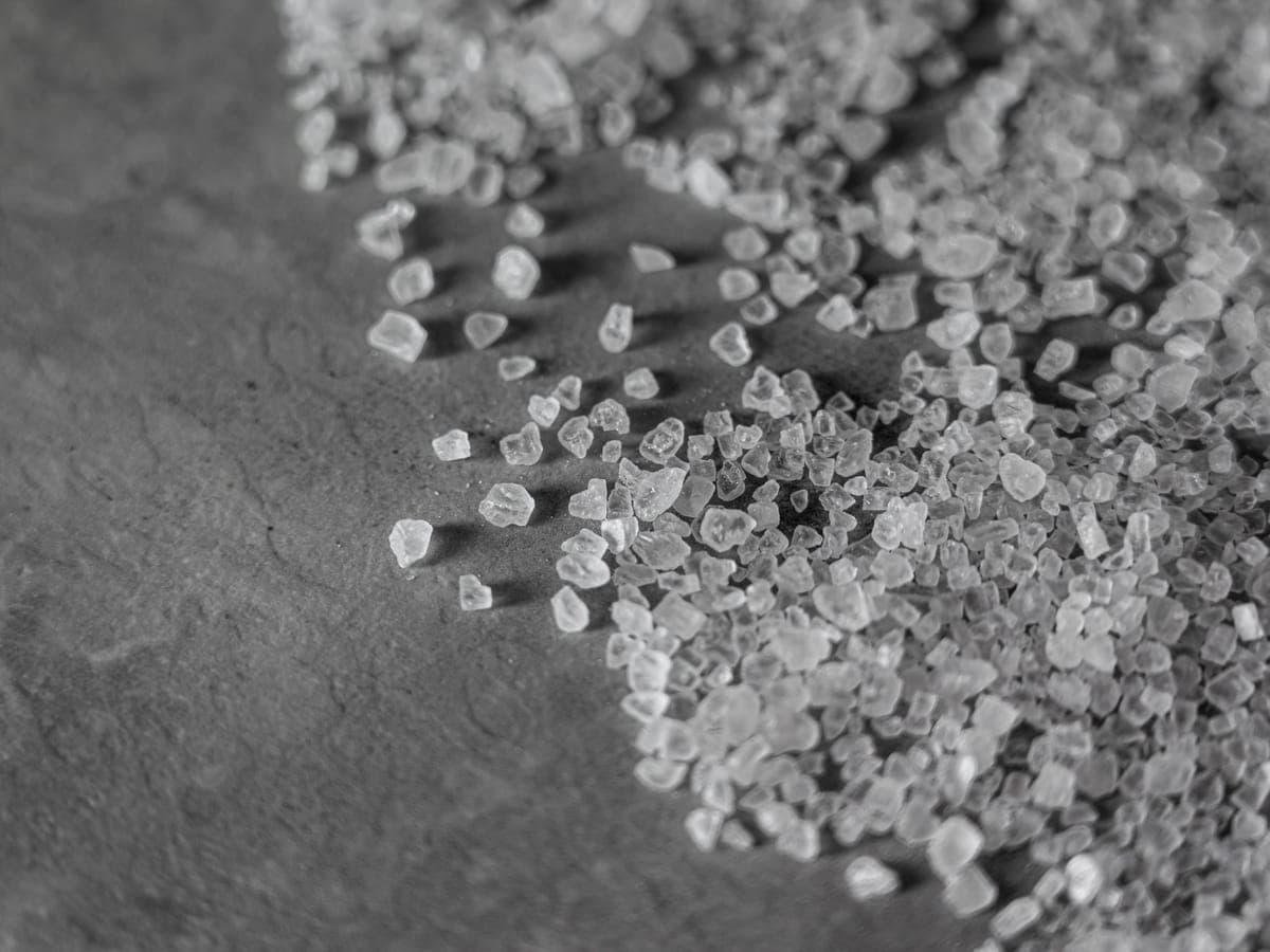 Primer plano de gruesos cristales de sal de roca esparcidos sobre una superficie oscura — el material granular que se esparce para deshelar el pavimento cubierto de nieve y hielo.