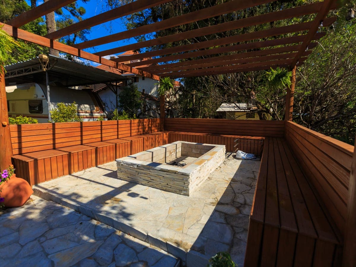 Una fogata rectangular de obra revestida de piedra sobre un patio de lajas, rodeada de bancas de madera fijas bajo una pérgola de madera.