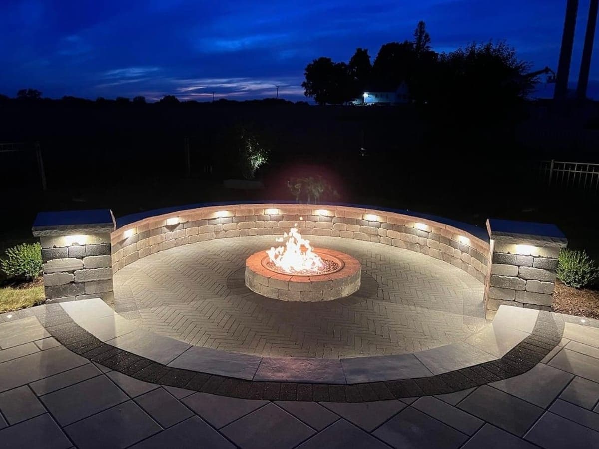 Gas fire pit burning at dusk inside a circular paver patio, surrounded by a curved seating wall with built-in lighting and two stone pillars.
