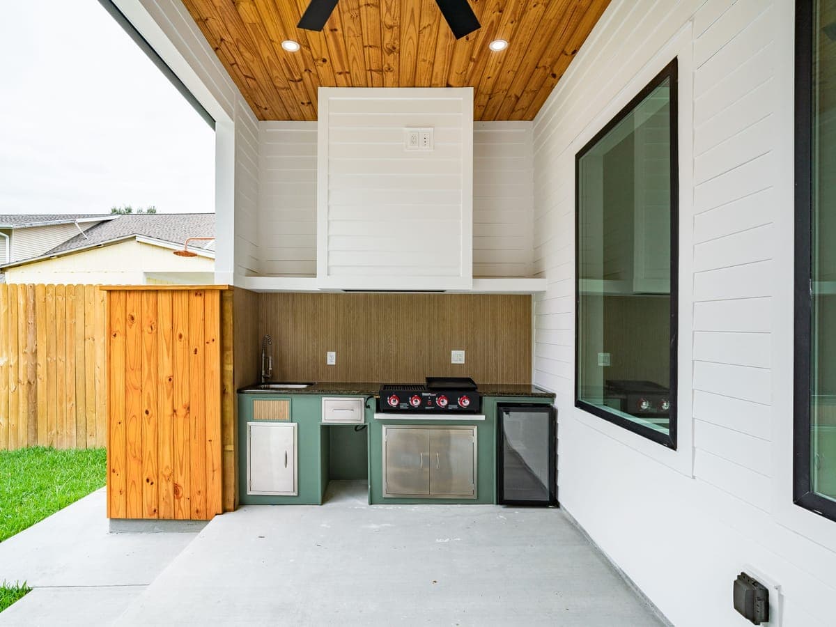 Una cocina exterior empotrada con parrilla de acero inoxidable, fregadero y refrigerador de bebidas integrados en los gabinetes, bajo un patio techado revestido de madera, junto a un césped cercado.