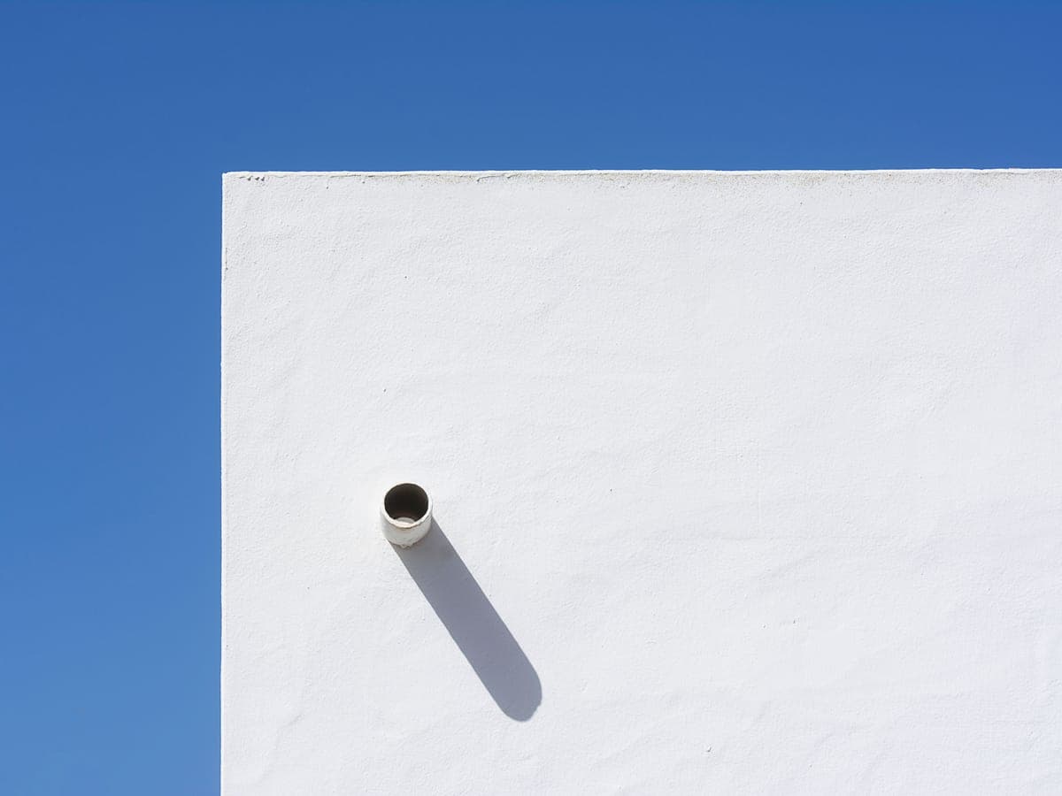 Un tubo de ventilación de PVC gris blanquecino que sobresale de la pared exterior de estuco blanco de un edificio contra un cielo azul despejado.