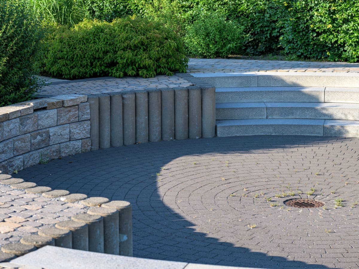 Un muro de contención curvo de bloques segmentados con remate de albardilla y escalones de piedra que rodea un patio circular de adoquines, bordeado de arbustos y setos.
