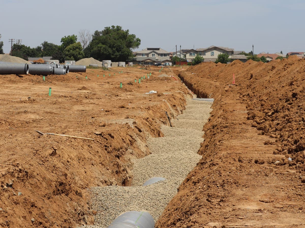 An open utility trench excavated across a construction site with drainage pipe staged alongside, before backfilling.