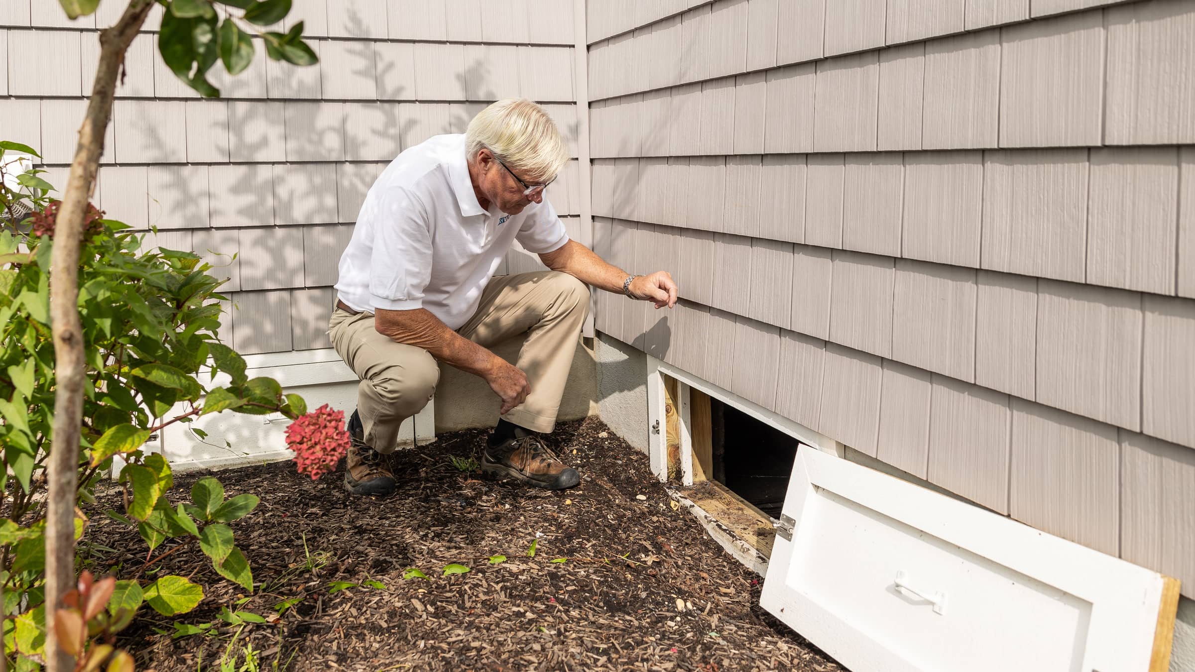 An open crawl-space access hatch at the base of a home's shingled exterior wall, with a person crouched beside the opening.