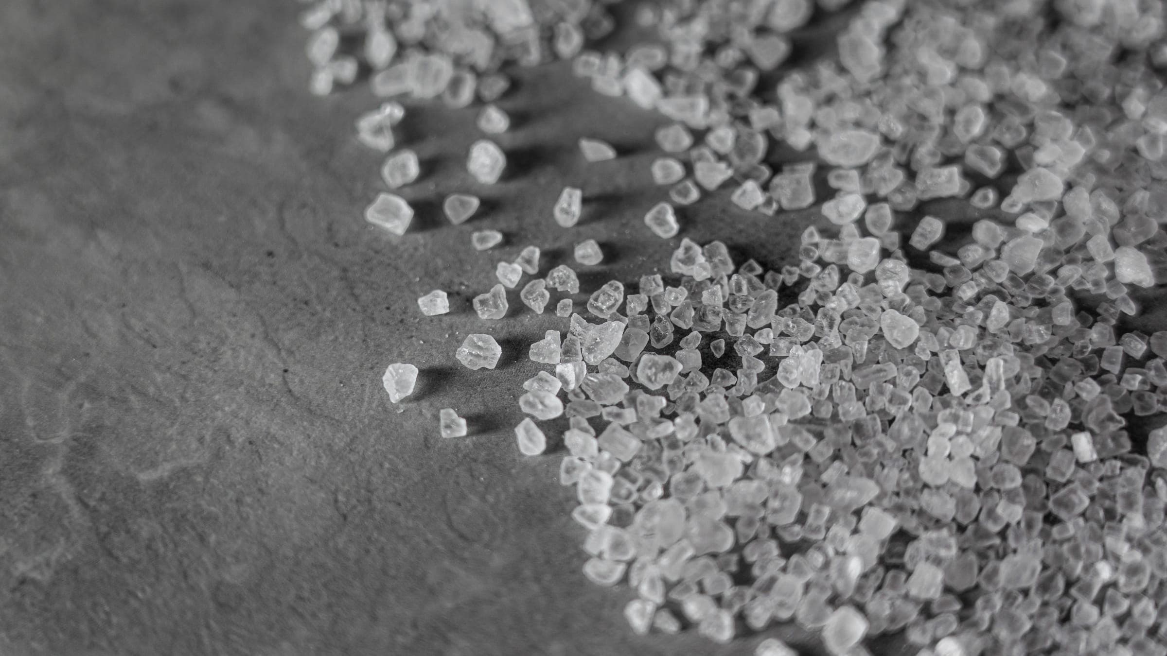 Primer plano de gruesos cristales de sal de roca esparcidos sobre una superficie oscura — el material granular que se esparce para deshelar el pavimento cubierto de nieve y hielo.