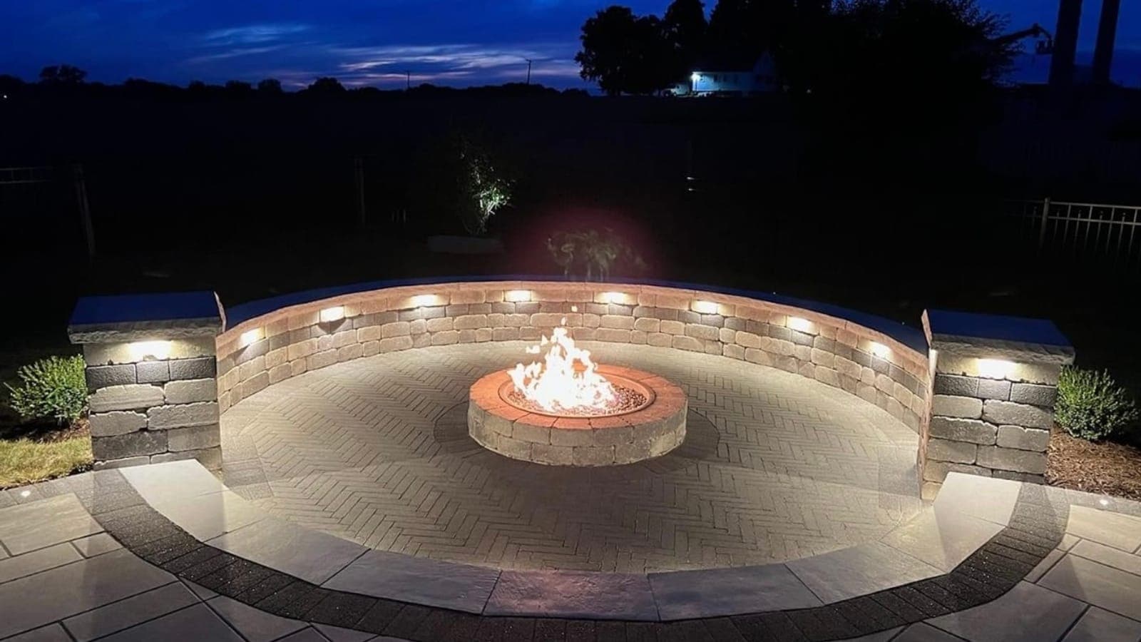 Gas fire pit burning at dusk inside a circular paver patio, surrounded by a curved seating wall with built-in lighting and two stone pillars.
