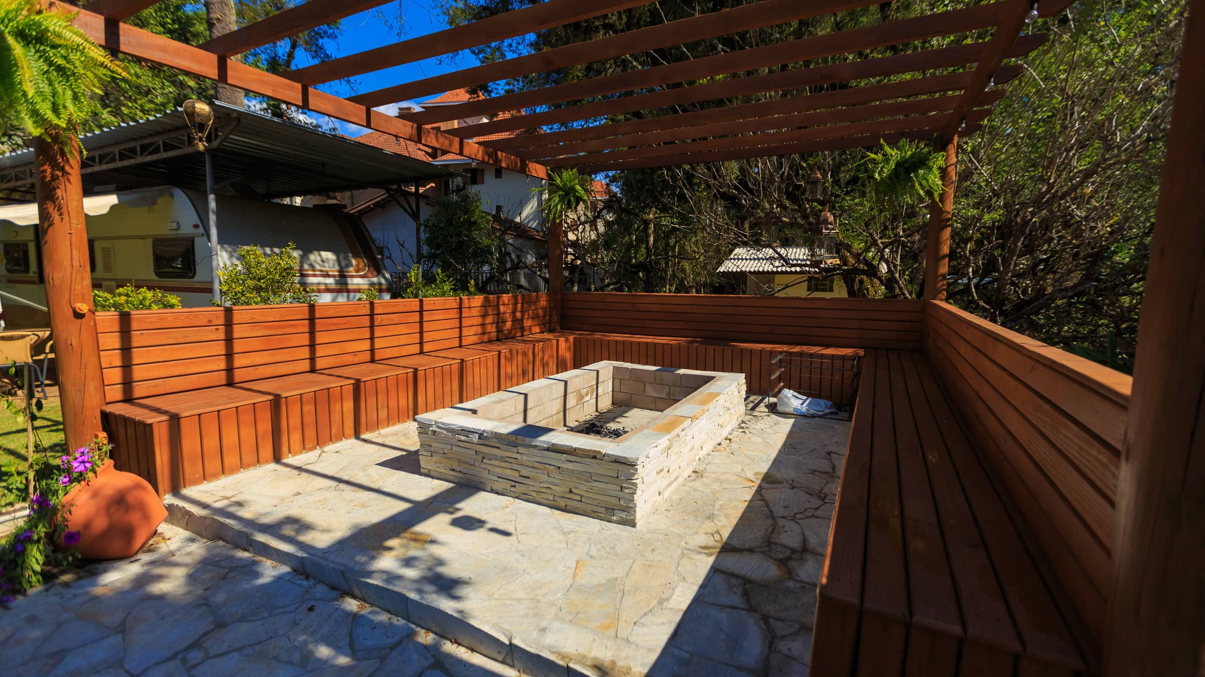 A built stone-clad rectangular fire pit on a flagstone patio, ringed by fixed wooden bench seating under a timber pergola.
