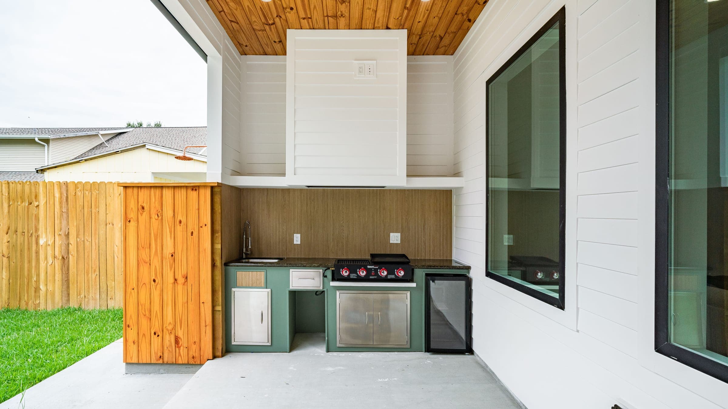 A built-in outdoor kitchen with a stainless grill, sink, and beverage fridge set into cabinetry under a wood-clad covered patio, beside a fenced lawn.