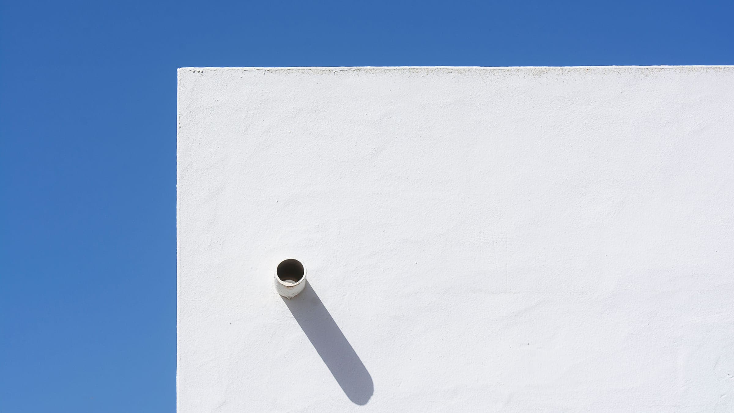 Un tubo de ventilación de PVC gris blanquecino que sobresale de la pared exterior de estuco blanco de un edificio contra un cielo azul despejado.