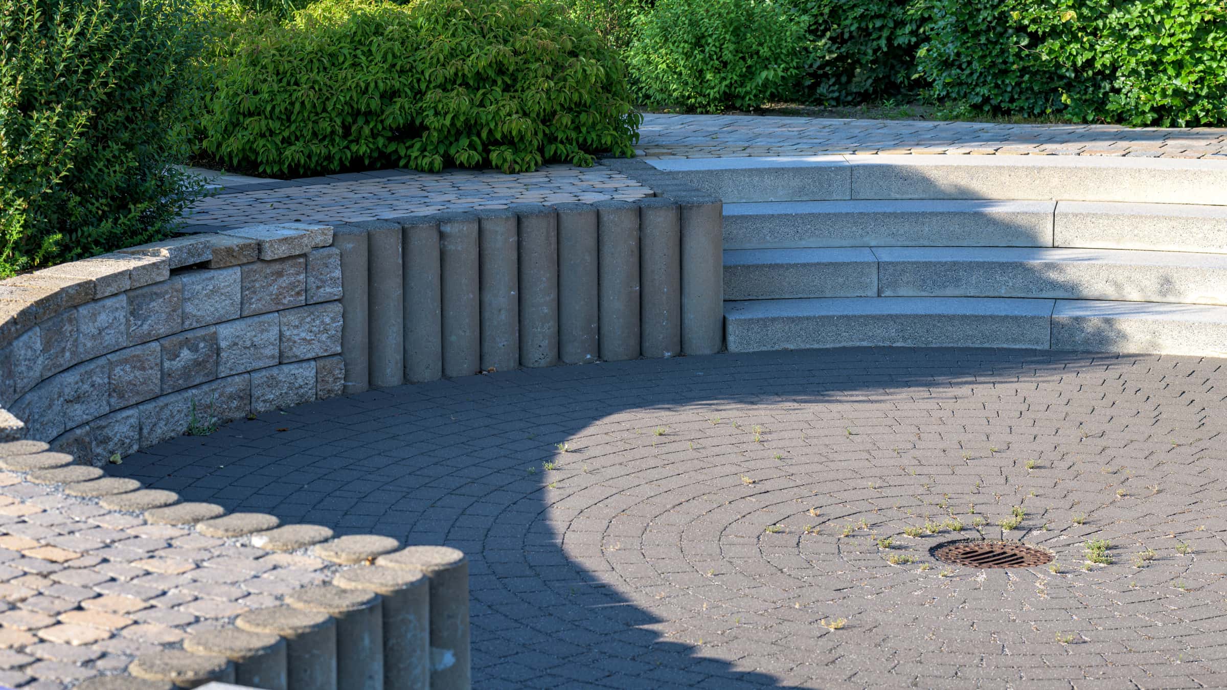 Un muro de contención curvo de bloques segmentados con remate de albardilla y escalones de piedra que rodea un patio circular de adoquines, bordeado de arbustos y setos.
