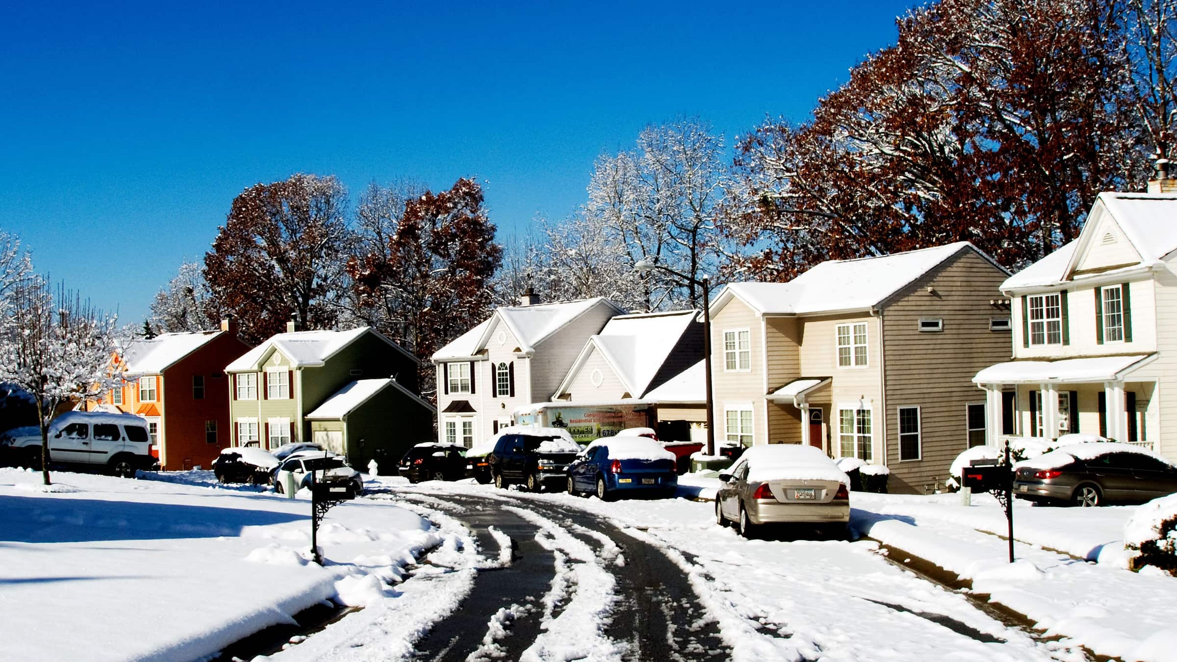 Una calle residencial suburbana cubierta de nieve, bordeada de casas y autos estacionados tras una nevada invernal.