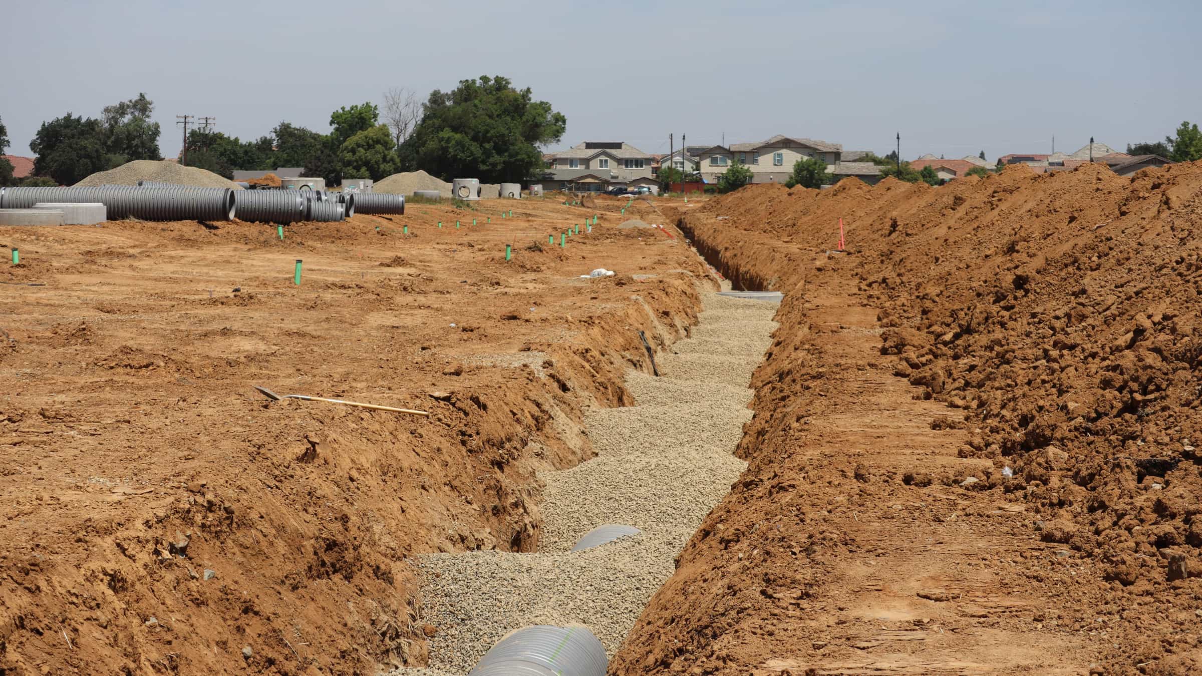 An open utility trench excavated across a construction site with drainage pipe staged alongside, before backfilling.